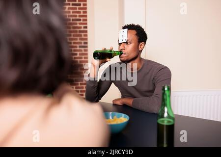 Selbstbewusster junger Erwachsener mit Plastikkarte auf der Stirn, der Bier trinkt, während er mit Freunden Charaden spielt. Person, die am Tisch im Wohnzimmer sitzt, während sie Spaß an Freizeitaktivitäten genießt. Stockfoto