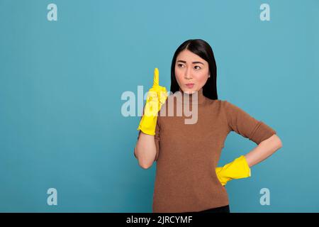 Junge asiatische Frau mit Verachtung Stimmung von Aufgaben tragen gelbe Handschuhe für die Handsicherheit und zeigen Finger nach oben Blick zur Seite isoliert auf einem blauen Hintergrund, Reinigung Home Konzept, Stockfoto