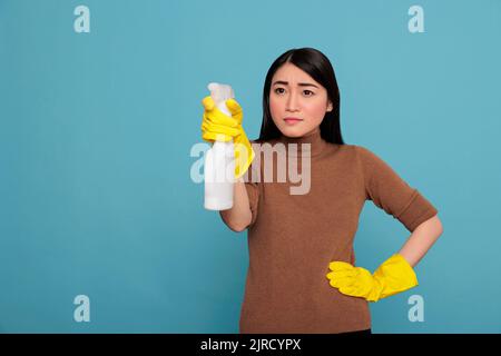 Vertrauen zufrieden asiatische junge Frau hält Spray in gelben Handschuh bereit für den Dienst vor einem blauen Hintergrund, Reinigung Home-Konzept, seriöse treue Frau von Tag zu Tag Hausarbeiten Stockfoto