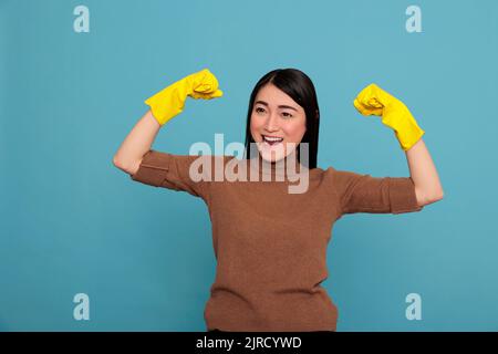 Aufgeregt glücklich und lächelnd asiatische Housekeeping heben die Arme und zeigen Bizeps isoliert blauen Hintergrund, Hausfrau Arbeiter, Reinigung Haus Konzept, energisch fröhlich und Lichtung mit positiven Geisteszustand Stockfoto