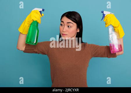 Traurige müde asiatische Hausfrau hob die Arme mit zwei Waschmittelsprays in den gelben Handschuhen vor blauem Hintergrund, Reinigung Hauskonzept, überarbeitete Hausfrau von Tag zu Tag Aufgaben Stockfoto