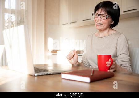 Ein glücklicher, anspruchsvoller Videoanruf einer älteren Frau auf einem Laptop, der von zu Hause aus läuft. Eine lächelnde Frau spricht Mitte 60s in einem virtuellen Online-Konferenzchat Stockfoto