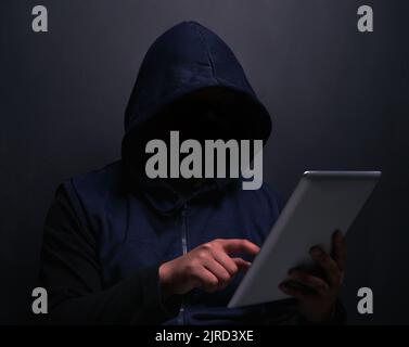 Wer sollte mein nächstes Opfer sein. Ein nicht erkennbarer Hacker, der ein digitales Tablet vor dunklem Hintergrund nutzt. Stockfoto