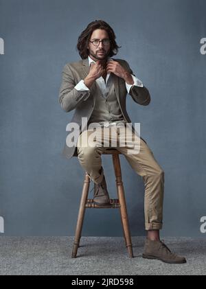 Ich bin ein Mann mit Stil. Ganzkörperportrait eines hübschen und stilvollen jungen Mannes, der auf einem Hocker vor einem grauen Hintergrund sitzt. Stockfoto