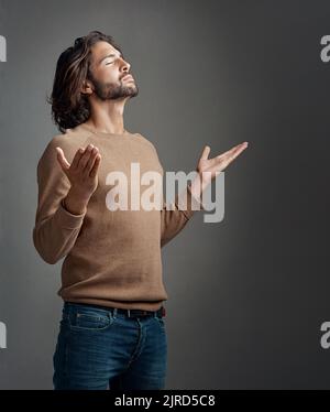 Lobend seine Anwesenheit. Studioaufnahme eines hübschen jungen Mannes, der vor grauem Hintergrund betet. Stockfoto