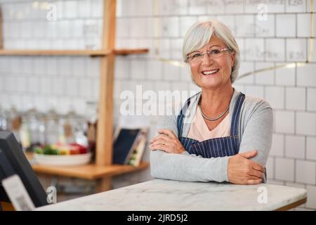 Mein Coffee Shop ist mein glücklicher Ort. Porträt einer glücklichen älteren Geschäftsbesitzerin, die an der Theke in ihrem Café posiert. Stockfoto