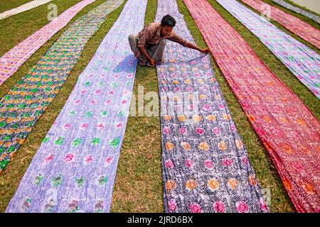 Narayanganj, Dhaka, Bangladesch. 23. August 2022. Farbenfrohe Stoffstreifen bilden ein auffälliges Display, da sie in ordentlichen Reihen auf einem Feld in Narayanganj, Bangladesch, angeordnet sind. Die langen Baumwolltücher werden lokal als ''Saree'' bezeichnet - ein traditionelles Kleidungsstück für Frauen, das unter der heißen Sonne trocknen soll und mit hellen Farben gefärbt wurde. Hier werden täglich etwa 4000 Stück Stoff zum Trocknen gelegt. Der Prozess dauert in der Regel drei Stunden, wobei jeder Satz von 200 Stück auf einmal bei Temperaturen trocknen kann, die über 36 Grad celsius erreichen können. Schön verzierte bunte lange Tücher sind c Stockfoto