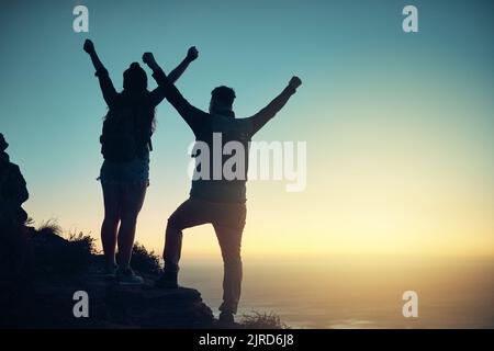 Wir haben es geschafft. Durchgehende Silhouette eines jungen Paares, das vom Gipfel des Berges aus feiert. Stockfoto