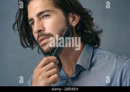Den bärtigen Look lieben. Studioaufnahme eines hübschen jungen Mannes, der seinen Bart vor einem grauen Hintergrund kämmt. Stockfoto