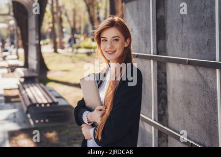 Eine ginger Geschäftsfrau genießt morgens ihren Weg ins Büro und hält ihren Laptop in den Händen. Unternehmensporträt. Schönheitsporträt. Stockfoto