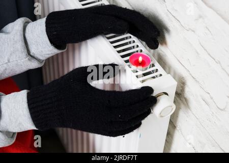 Der Besitzer wärmt sich am Heizkörper in warmen Fäustlingen auf. Harter Winter. Heizungskrise Stockfoto