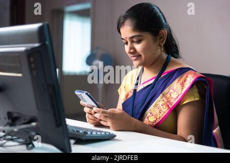 Lächelnder Bankangestellter, der im Büro vor dem Computer mit dem Mobiltelefon telefoniert - Konzept der Entspannung, des Kundenservice und der beruflichen Tätigkeit Stockfoto