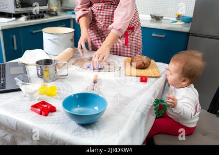Eine Mutter macht in der Küche Kekse. Das Baby sitzt auf einem Stuhl und beobachtet den Prozess. Auf dem Tisch befindet sich ein Laptop. Nahaufnahme. Draufsicht. Das c Stockfoto