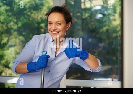 Hausfrau in Gummihandschuhen, lächelnd, Daumen nach oben zeigend, Kamera anblickend, glücklich Hausarbeiten erledigen, Boden wischen Stockfoto