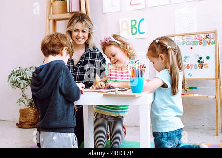 Porträt einer jungen Frau, die am Tisch um spielende Kinder sitzt. Lehrer beobachten Schüler, die Aufgaben im Klassenzimmer erledigen. Stockfoto