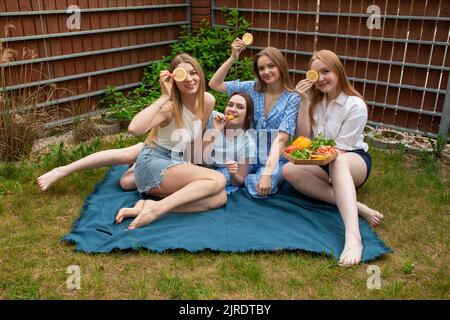 Portrait von jungen Frauen, die die Augen mit Orangenscheiben bedeckten, geschnittene Früchte auf dem Tablett hielten, auf dem karierten Garten sitzend. Stockfoto