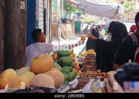 ESNA, LUXOR, ÄGYPTEN - 13. AUGUST 2022: Verschleierte Frauen in traditioneller schwarzer Kleidung kaufen Früchte auf dem lokalen Markt in der Stadt Esna, südlich von Luxor. Stockfoto