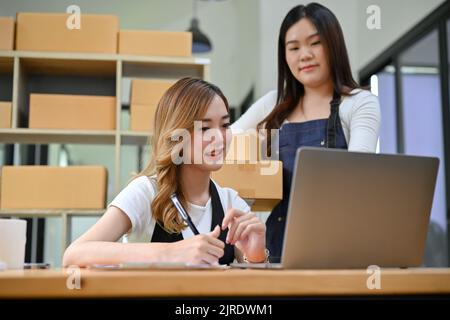 Zwei attraktive junge asiatische Unternehmerinnen arbeiten im Verpackungsraum zusammen, bereiten den Auftrag ihres Kunden vor, überprüfen die Bestellungen Stockfoto