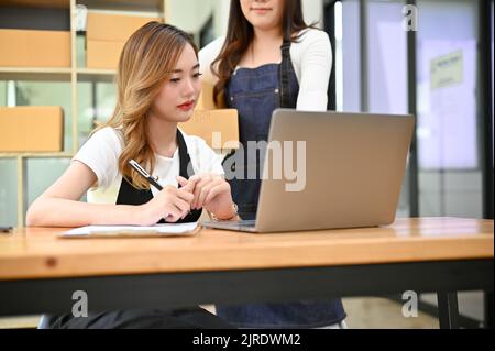Schöne junge asiatische weibliche Online-Shop-Besitzer arbeitet mit ihrem Geschäftspartner im Büro, überprüfen Versand und Lagerstatus auf Laptop-Computer. Stockfoto