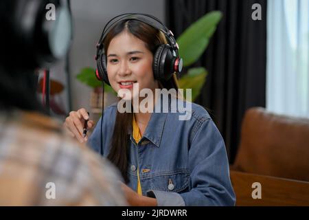 Die selbstbewusste junge asiatische Radiomoderatorin oder Podcast-Content-Creator zeichnet ihren Podcast auf und interviewt ihren Gast für ihre Online-Radioinhalte. Stockfoto