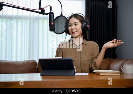 Selbstbewusste und schöne junge asiatische Radiomoderatorin, die Kopfhörer trägt, auf ihrem Mikrofon spricht und ihren Online-Radiosender aufnimmt Stockfoto