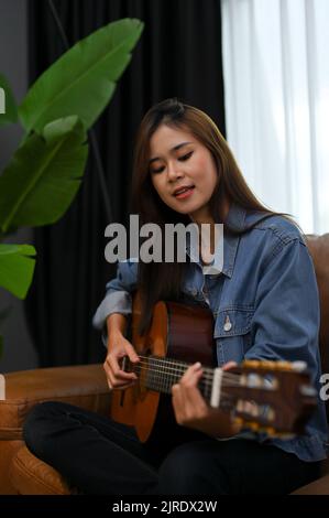 Schöne und talentierte asiatische Mädchen in jean Jacke singen, während sie ihre akustische Gitarre in ihrem modernen Wohnzimmer spielen. Stockfoto