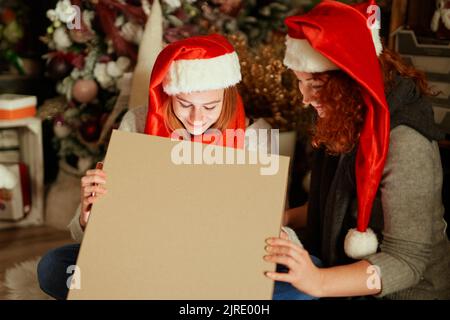 Weihnachtszeit Konzept. Tochter in rot Weihnachtsmann Hut öffnet Weihnachts-Geschenkbox, Mädchen, die in der Box mit Licht innen, Mama sitzt in der Nähe. Ausdruckskonzept. Hochwertige Bilder Stockfoto