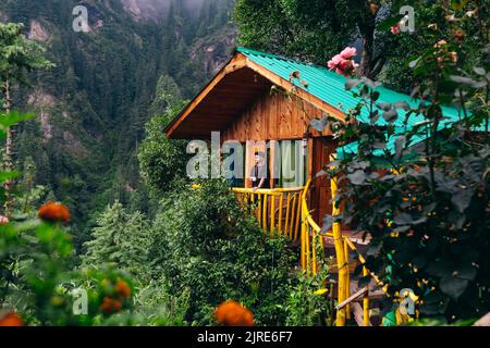 Mann auf dem Balkon eines hölzernen Baumhauses, versteckt im Zedernwald der Jibhi-Berge in Nordindien Stockfoto