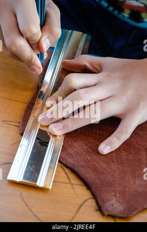 Nahaufnahme der Hände eines Teenagers mit einem scharfen Skalpell und einem Metalllineal als Anleitung zum Schneiden eines geraden Lederstücks. Stockfoto