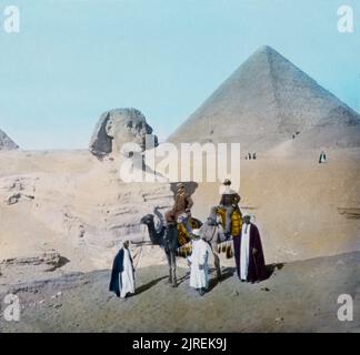 Ein Vintage handkoloriertes Dia-Foto mit magischer Laterne aus dem Jahr 1885 von viktorianischen Touristen auf Kamelen, die vor der Großen Sphinx von Gizeh in Ägypten posieren, mit der Pyramide von Khafre im Hintergrund des Fotografen William Henry Jackson Stockfoto