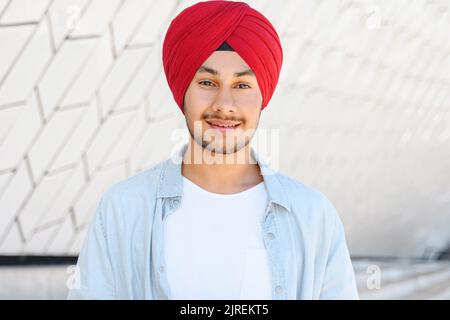 Junger hübscher indischer Mann in rotem, traditionellem Turban und lässigem Hemd, der im Freien steht, lächelnd, angenehmer hindu-Student, der die Kamera anschaut, Headshot Stockfoto