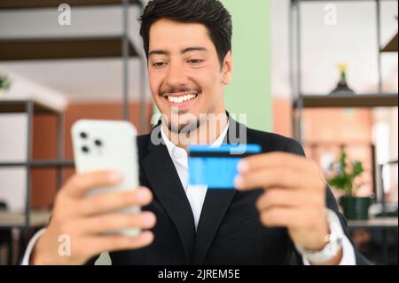 Fröhlicher lateinischer Mann macht online kaufen. Hispanic Kerl in Business Wear hält Telefon und eine Kreditkarte, macht Geldtransaktion online sitzen im Büro, Kerl ist mit Smartphone und Debit-Bankkarte Stockfoto