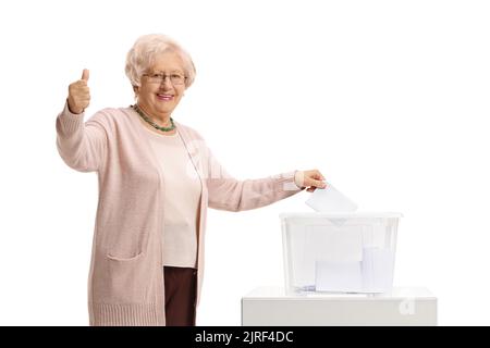 Eine ältere Dame, die in einer Wahlurne abstimmen und auf weißem Hintergrund die Daumen nach oben zeigen kann Stockfoto
