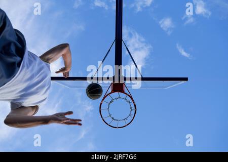 Der Mann trainiert auf dem Straßenplatz Basketball. Er wirft den Ball auf den Reifen. Niedriger Winkel Stockfoto