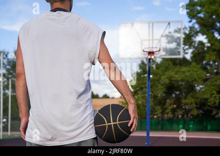 Der Mann steht mit dem Ball in der Hand auf dem Hintergrund des Basketballplatzes im Park. Street Basketball Stockfoto