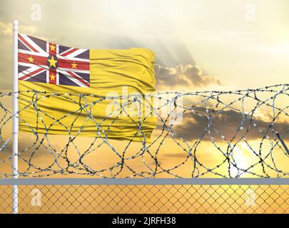 Fahnenmast mit der Fahne von Niue gegen den Himmel und hinter einem Zaun mit Stacheldraht. Das Konzept des Schutzes der Grenzen von Gebieten. Stockfoto