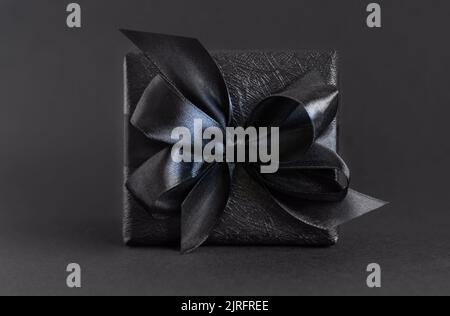 Schwarze Geschenkbox mit Schleife in Nahaufnahme auf Schwarz. Eingewickeltes Geschenk für Dark Motto Party, Weihnachten, Geburtstag, Jubiläum. Black Friday Konzept Stockfoto