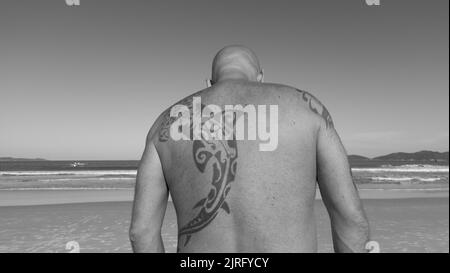 Bombinhas, Santa Catarina, Brasilien - 22. Mai 2022: Mann mit einem Hai-Tattoo auf dem Rücken, der an einem sonnigen Tag in Südbrasilien am Strand steht. Stockfoto