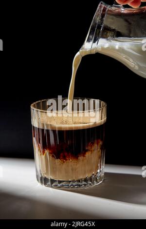 Hand mit Kanne Gießen von Creme auf ein Glas Kaffee Stockfoto