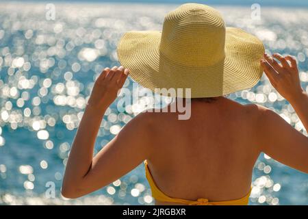 Junge Frau in einem gelben Bikini, die sich an ihrem Hut festhält und auf das Meer blickt. Stockfoto