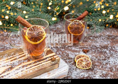 Zwei Tassen Tee aus Glas mit Zitrone, Zimtstangen auf einem Holztisch vor dem Hintergrund eines grünen Weihnachtsbaums mit Lichtergirlanden. Stockfoto