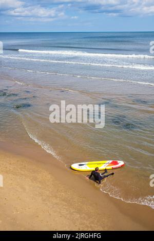 Ein Paddelmann in einem Neoprenanzug sitzt im flachen Wasser mit leerem Strand in North Bay, Scarborough Stockfoto