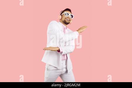 Fröhlicher, lustiger junger Mann in einem weißen Anzug, einem rosa Hemd und einer Sonnenbrille mit kabellosen pinken Kopfhörern hört Musik und tanzt in einem pinken Studio Stockfoto