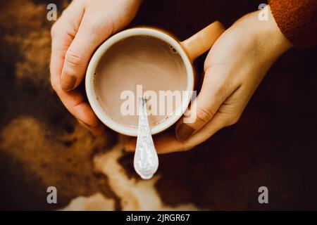 Draufsicht auf einen Becher warmen Kaffees mit Milch, der in den Händen einer Frau in einer braunen Jacke vor dem Hintergrund von Kaffeeflecken auf einem dunklen gehalten wird Stockfoto