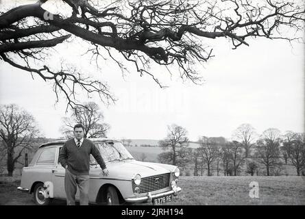 1958, historisch, draußen auf dem Land, auf einer kleinen Landstraße, ein Mann, der neben einem Austin A40 Farina-Auto der damaligen Zeit stand, England, Großbritannien. Es handelt sich um ein modernes kleines Auto mit einer markanten „zwei-Kasten“-Form und Kopffreiheit auf dem Rücksitz, das von dem berühmten italienischen Automobildesigner Battista Farina entworfen wurde, der für seine Arbeit mit Ferrari und dem Alfa Romeo Spider berühmt ist. Das Auto wurde bis 1967 produziert, als es vom Austin 1100 abgelöst wurde. Stockfoto