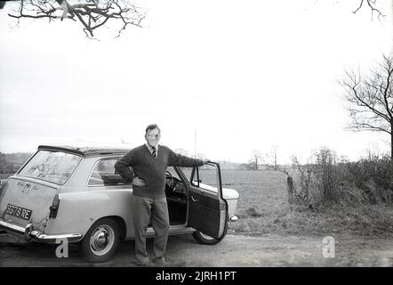 1958, historisch, draußen auf dem Land, auf einer kleinen Landstraße, ein Mann, der neben einem Austin A40 Farina-Auto der damaligen Zeit stand, England, Großbritannien. Es handelt sich um ein modernes kleines Auto mit einer markanten „zwei-Kasten“-Form und Kopffreiheit auf dem Rücksitz, das von dem berühmten italienischen Automobildesigner Battista Farina entworfen wurde, der für seine Arbeit mit Ferrari und dem Alfa Romeo Spider berühmt ist. Das Auto wurde bis 1967 produziert, als es vom Austin 1100 abgelöst wurde. Stockfoto