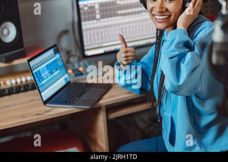 Nahaufnahme einer hübschen afrikanischen Anchorfrau mit Kopfhörern, die den Daumen nach oben zeigen, während sie im Aufnahmestudio im Radio ausgestrahlt wird Stockfoto
