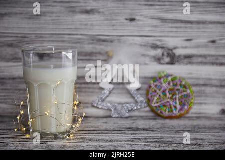 Süße Weihnachtstradition. Handgemachte frische Kekse und Glas mit Milch für den Weihnachtsmann auf dem Vintage-Holztisch. Festliche Weihnachtsstimmung und ästhetische Atmosphäre mit Girlanden Hintergrund. Speicherplatz kopieren. Stockfoto