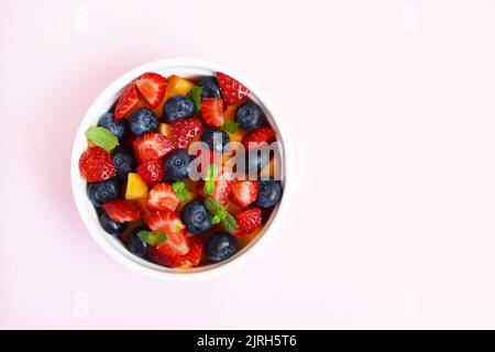 Obstsalat in einer weißen Tasse auf einem weichen rosa Hintergrund. Erdbeeren, Pfirsiche und Heidelbeeren in Scheiben geschnitten mit Minzblättern. Freier Speicherplatz für Text. Stockfoto