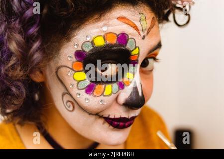 Nahaufnahme der jungen kaukasischen lateinerin, die lächelt und mit dem Make-up von La Calavera Catrina in ihrem Zimmer auf die Kamera schaut und sich auf das Feiern vorbereitet Stockfoto